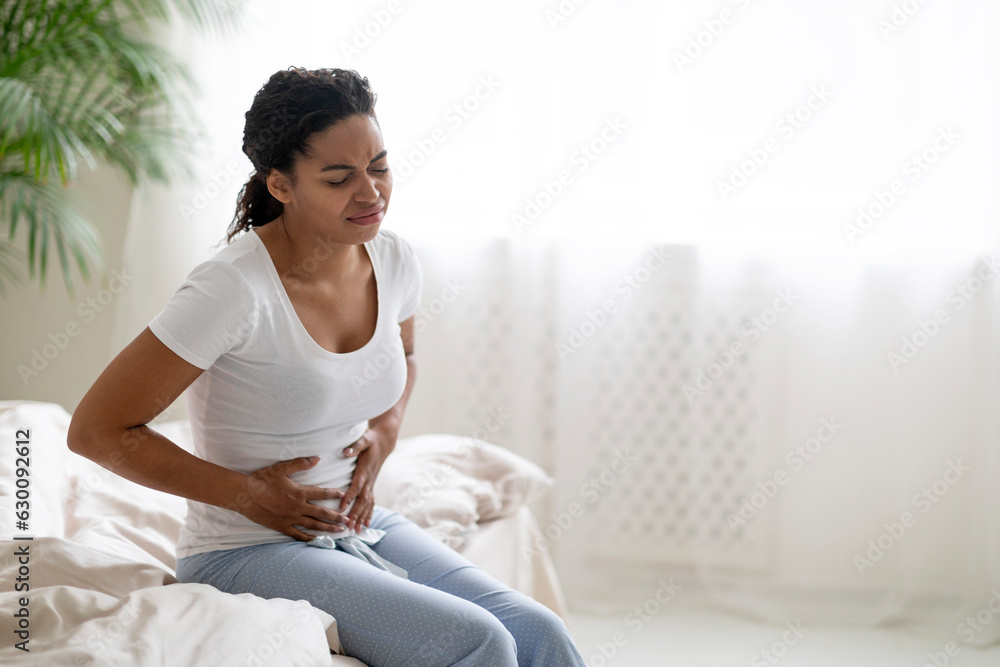 Black woman suffering stomach pain, sitting on bed and touching belly Black woman suffering stomach pain, sitting on bed and touching belly