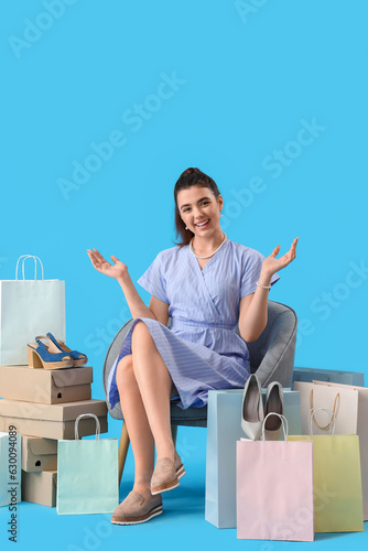 Young woman with shoe boxes and shopping bags sitting in armchair on blue background