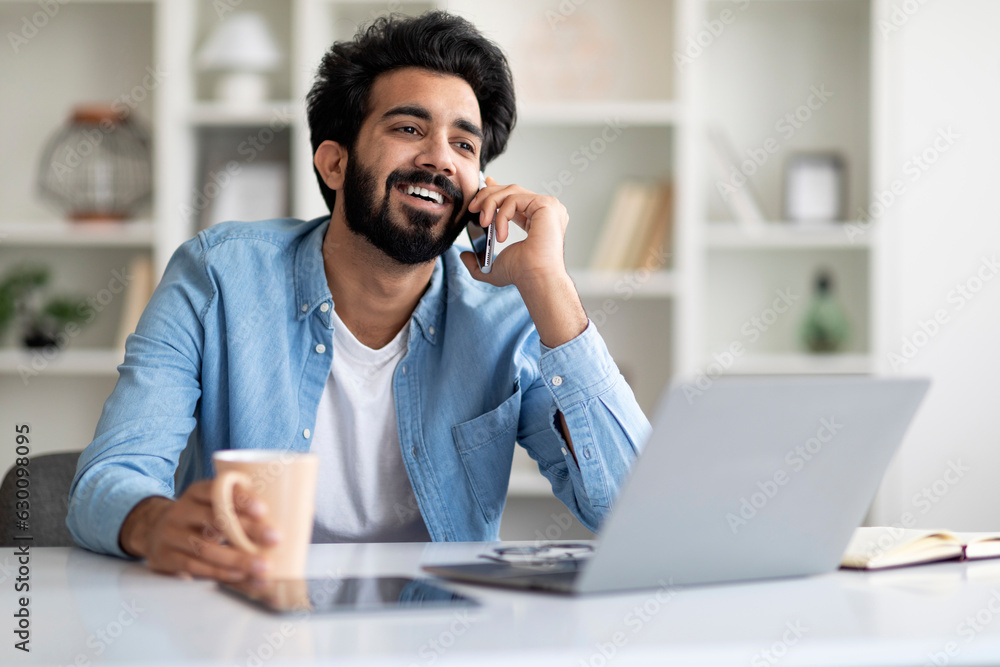 Cheerful Indian Freelancer Guy Drinking Coffee And Talking On Cellphone At Home Cheerful Indian Freelancer Guy Drinking Coffee And Talking On Cellphone At Home