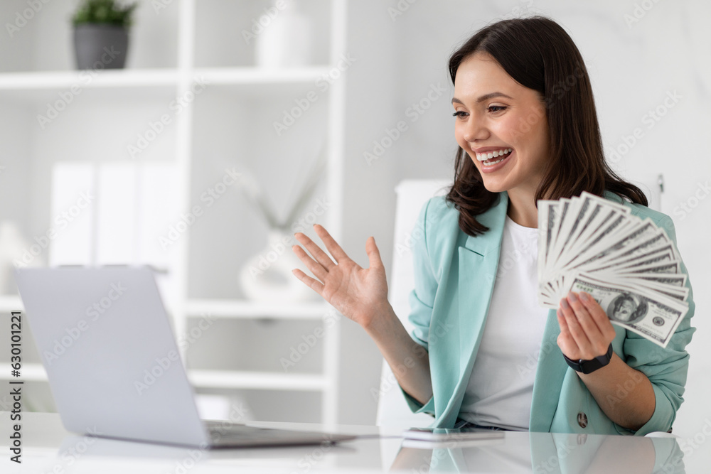 Cheerful excited shocked young caucasian businesswoman in suit at table with computer hold lot of money dollars Cheerful excited shocked young caucasian businesswoman in suit at table with computer hold lot of money dollars
