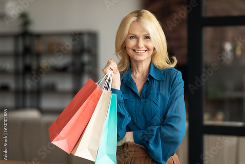 Portrait of happy mature woman holding bright shopping bags and smiling at camera, posing in home interior