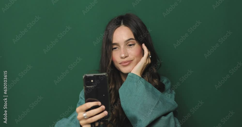 Happy girl stares at the phone and make photo. Female uses phone like mirror. Young girl make photo from hands with phone on green background. Curly girl wear green dress, smiling and look happy. Happy girl stares at the phone and make photo. Female uses phone like mirror. Young girl make photo from hands with phone on green background. Curly girl wear green dress, smiling and look happy.
