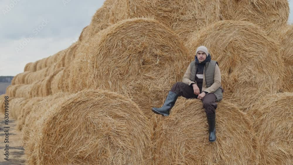 Long shot of modern Caucasian male farmer spending time outdoors sitting on haystack on cold autumn day Long shot of modern Caucasian male farmer spending time outdoors sitting on haystack on cold autumn day