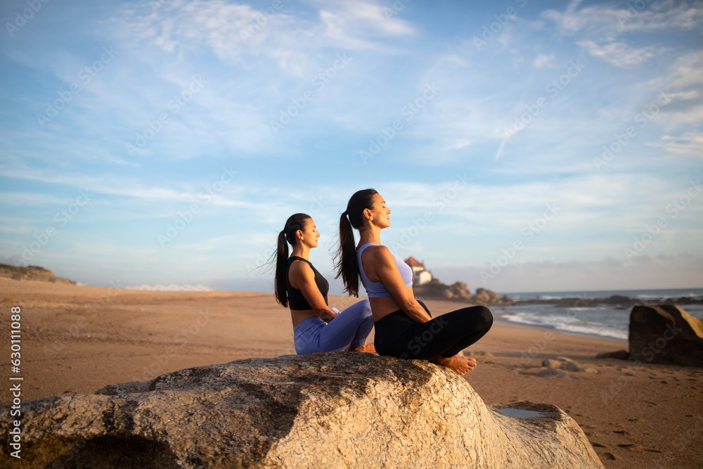 Cheerful millennial caucasian sisters women practice yoga with closed eyes enjoy breathing exercises, sunrise on beach