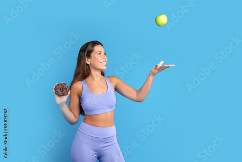Smiling slim young caucasian woman in sportswear with donut in hand throws up green apple, has fun © Prostock-studio
