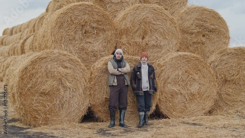 Long shot of confident man and woman working at farm standing against haystacks on cold autumn day looking at cameraLong shot of confident man and woman working at farm standing against haystacks on c photo