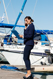 Female yachting dealer in marine blue suit holding notepad, standing outside a luxury sailboat using cell phone, Heraklion, Crete, Greece