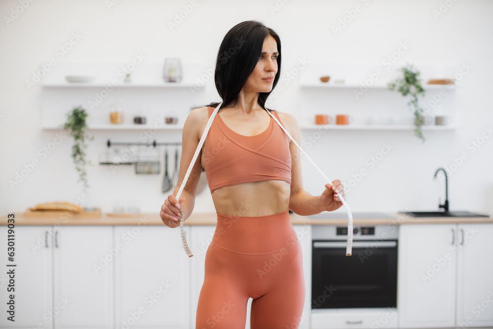 Slender lady in activewear stretching measuring tape on shoulders while looking into distance in spacious apartment. Pretty caucasian sportswoman staying motivated for leading healthy lifestyle. Slender lady in activewear stretching measuring tape on shoulders while looking into distance in spacious apartment. Pretty caucasian sportswoman staying motivated for leading healthy lifestyle.