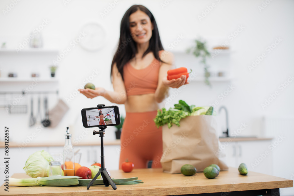 Selective focus of cell phone on tripod being used by cheerful lady with fresh salad ingredients from shopping bag. Fitness and nutrition blogger sharing tips on choosing proper products for meals.