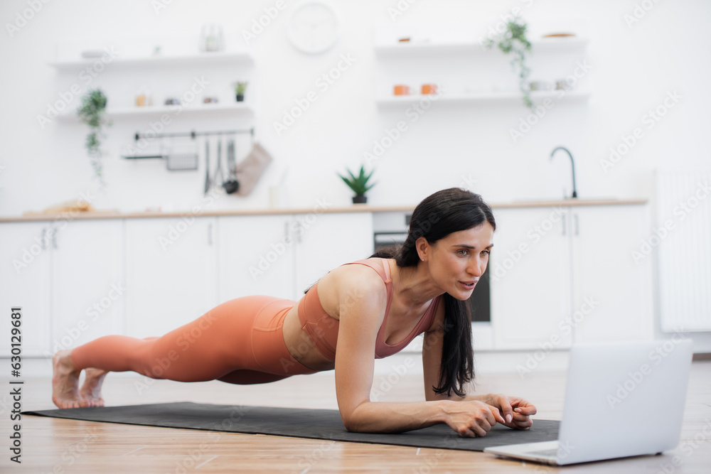 Strong female person standing in plank position and watching motivational video with online training on remote gadget. Flexible lady strengthening core and abs muscles while exercising on rug indoors. Strong female person standing in plank position and watching motivational video with online training on remote gadget. Flexible lady strengthening core and abs muscles while exercising on rug indoors.