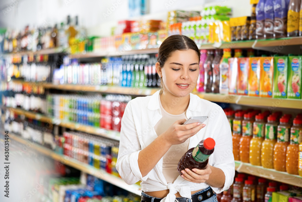Portrait of modern young girl scanning barcode on bottle of fruit juice with smartphone, paying for item using mobile app while shopping in supermarket..