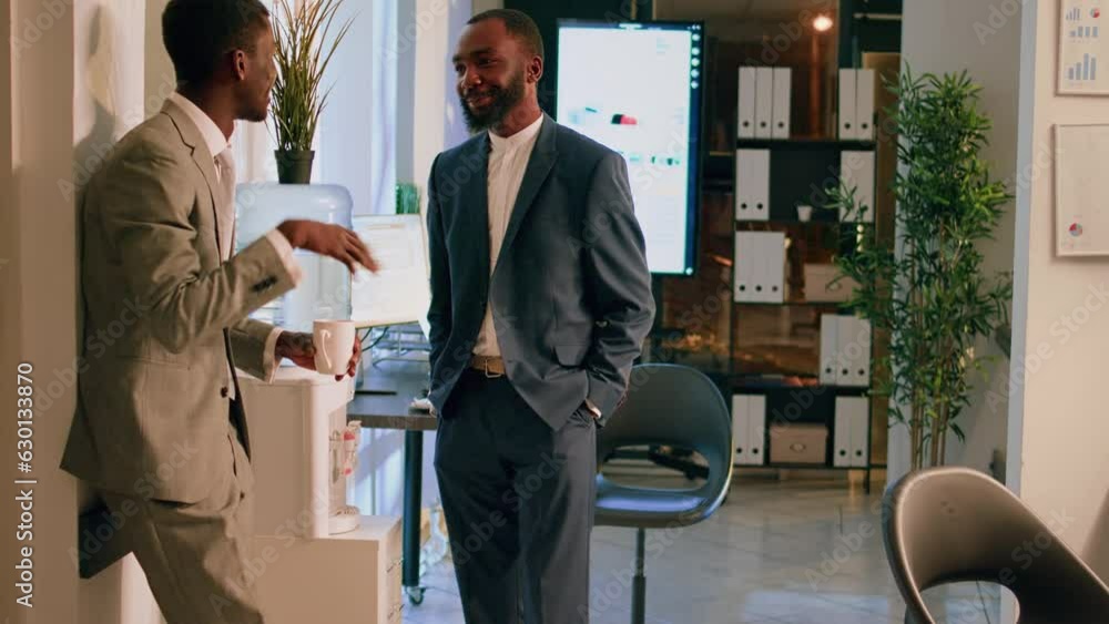 Relaxed coworking businessmen having pleasant discussion by water cooler in office overnight. Staff personnel enjoying cup of coffee together, chatting during nightshift break