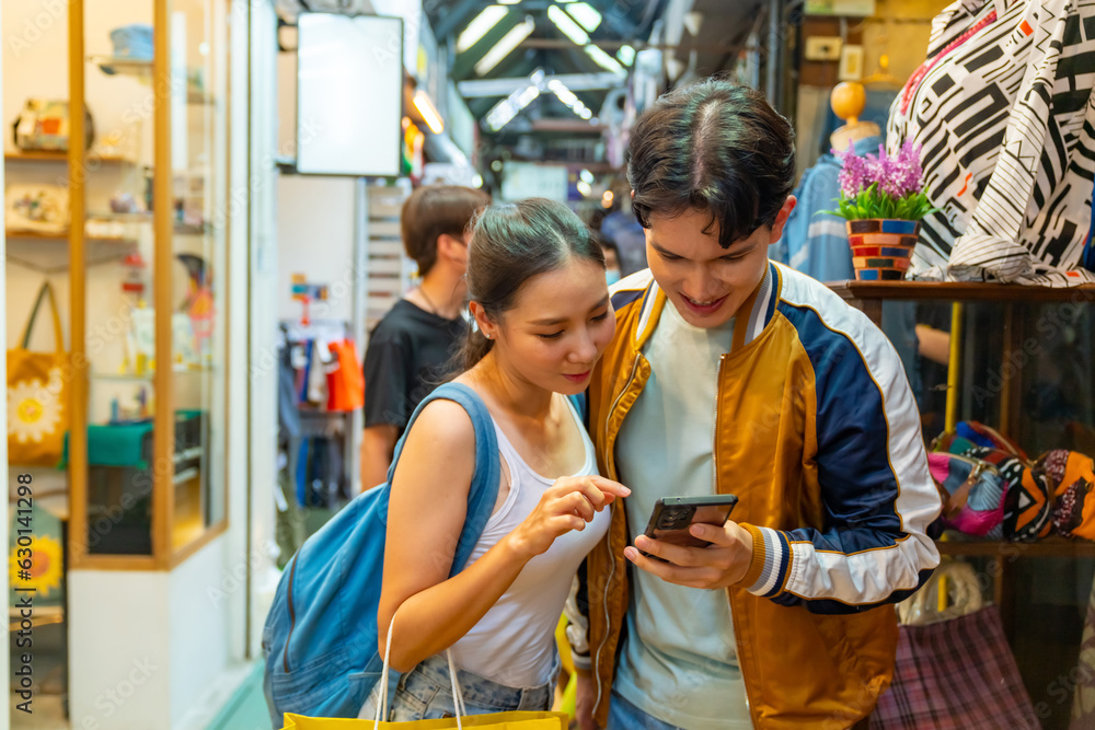 Asian couple enjoy and fun outdoor lifestyle in the city using mobile phone booking hotel during shopping together at chatuchak weekend street market in Bangkok, Thailand on summer holiday vacation.