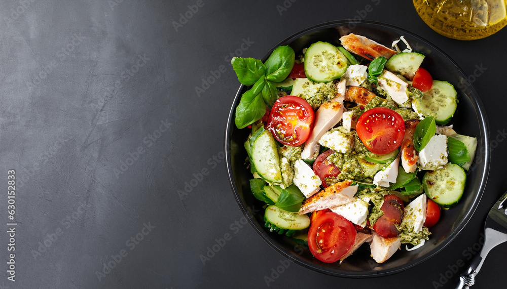 Tasty appetizing fresh salad with chicken, tomatoes, cucumbers, pesto sauce and cheese feta in bowl on black background. View from above. Horizontal with copy space. Tasty appetizing fresh salad with chicken, tomatoes, cucumbers, pesto sauce and cheese feta in bowl on black background. View from above. Horizontal with copy space.