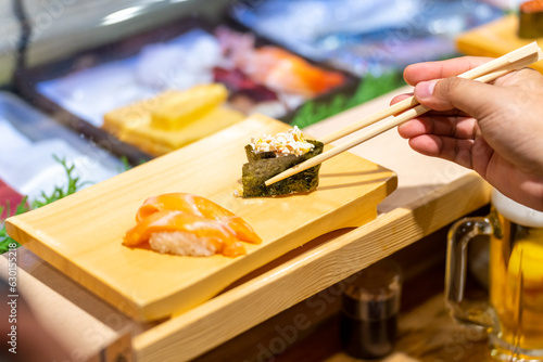 Asian woman hand using chopsticks eating Japanese food nigiri sushi with seaweed on sushi board on open kitchen counter. Chef serving assorted sushi and sashimi to customer at Japanese restaurant.