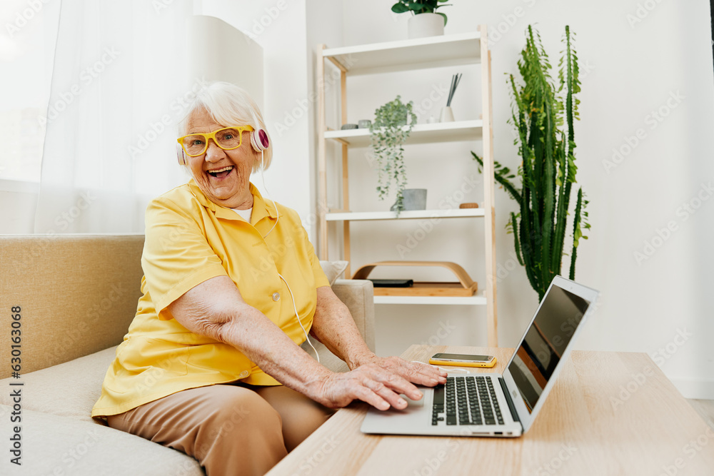 Happy elderly woman with a laptop typing with headphones sitting at home on the couch in a yellow shirt, bright modern interior, lifestyle online communication. Happy elderly woman with a laptop typing with headphones sitting at home on the couch in a yellow shirt, bright modern interior, lifestyle online communication.