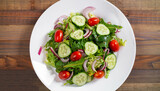 Salad with greens, cherry tomatoes, red onions and cucumber on white plate with wood background, overhead view