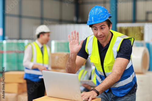 Architect or engineering man and worker standing and checking large warehouse with computer. Caucasian business manager raising hand and say hi with warehouse building background.