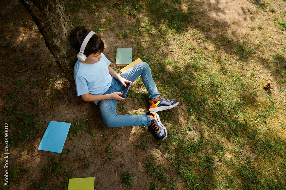 Fototapeta premium Schoolboy in wireless headphones, top view, playing online video games. Kids Education Digital addiction Entertainment