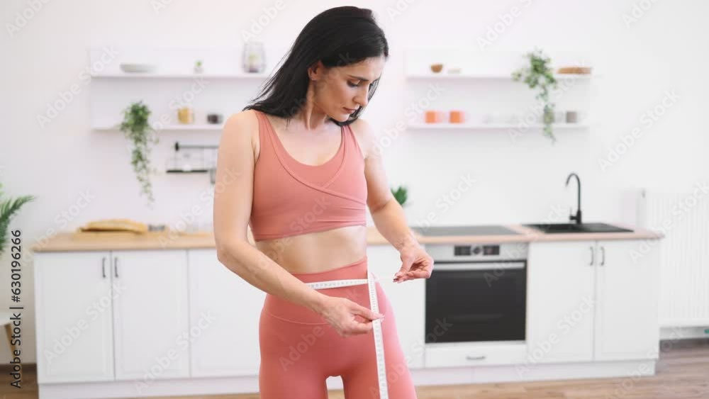 Attractive woman in good health measuring waist with tape after working out at home in yoga activewear. Athletic person steering in direction of improving well-being via food habits and fitness. Attractive woman in good health measuring waist with tape after working out at home in yoga activewear. Athletic person steering in direction of improving well-being via food habits and fitness.