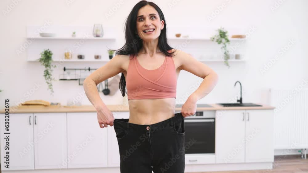 Surprised lady in terracotta top looking at black oversized jeans on thin waist while standing in kitchen of apartment. Sporty slim woman enjoying amazing achievements of well-balanced diet at home. Surprised lady in terracotta top looking at black oversized jeans on thin waist while standing in kitchen of apartment. Sporty slim woman enjoying amazing achievements of well-balanced diet at home.