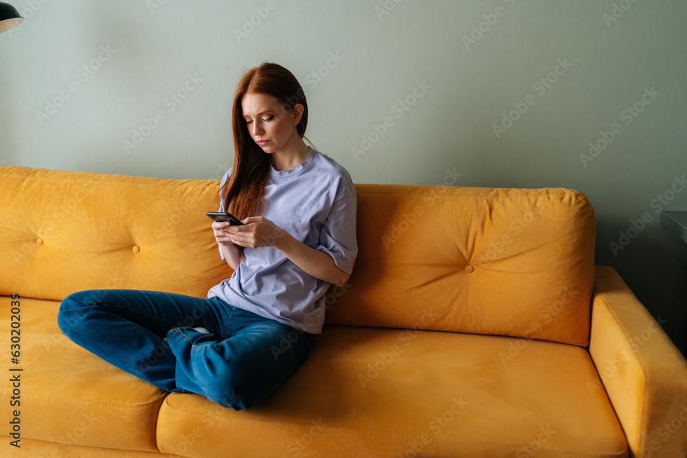 Portrait of frustrated young woman sitting on yellow couch looking at smartphone screen thinking about problem difficulty, feeling failure and exhausted, suffering grief and bad relationship Portrait of frustrated young woman sitting on yellow couch looking at smartphone screen thinking about problem difficulty, feeling failure and exhausted, suffering grief and bad relationship