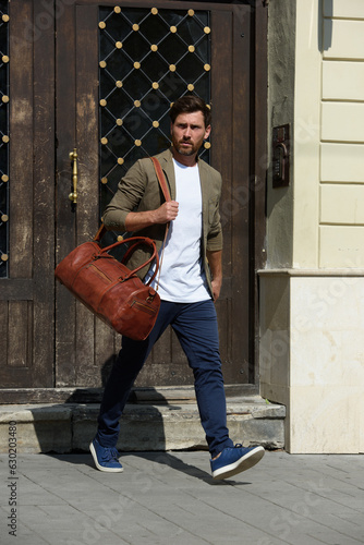 a man with a bag. black leather travel bag, street photo. man is wearing blue sneakers, jeans, a white T-shirt and a jacket