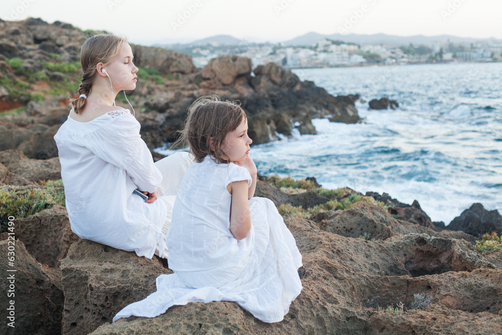 Two sisters in white clothes, kid and teenage girl sitting together near sea