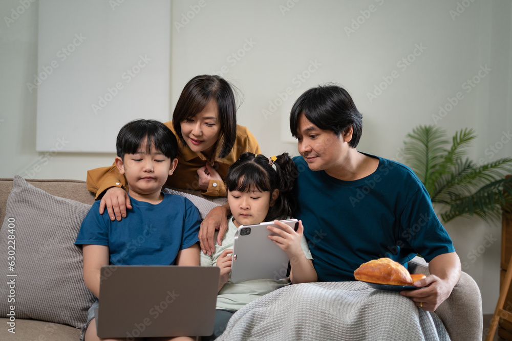 Happy parents with two kids using smartphone at home together, sitting on couch, smiling mother and father with little son and daughter looking at phone screen, having fun