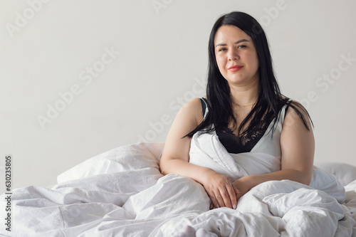 Black-haired woman wakes up in great mood in cozy bedroom. Adult housewife with long hair doing light stretch sits on white double bed, copyspace