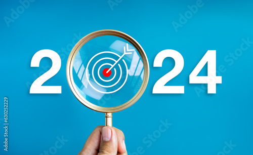 Happy new year 2024 with business concept banner. The big white 2024 year number with target dart icon inside the golden magnifying glass in hand on blue background. Trends, plan and goals concepts.