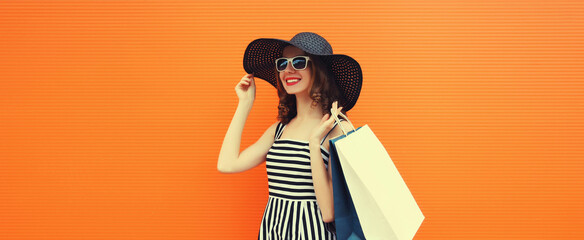 Happy smiling young woman with shopping bags wearing black summer straw hat, striped dress on orange background