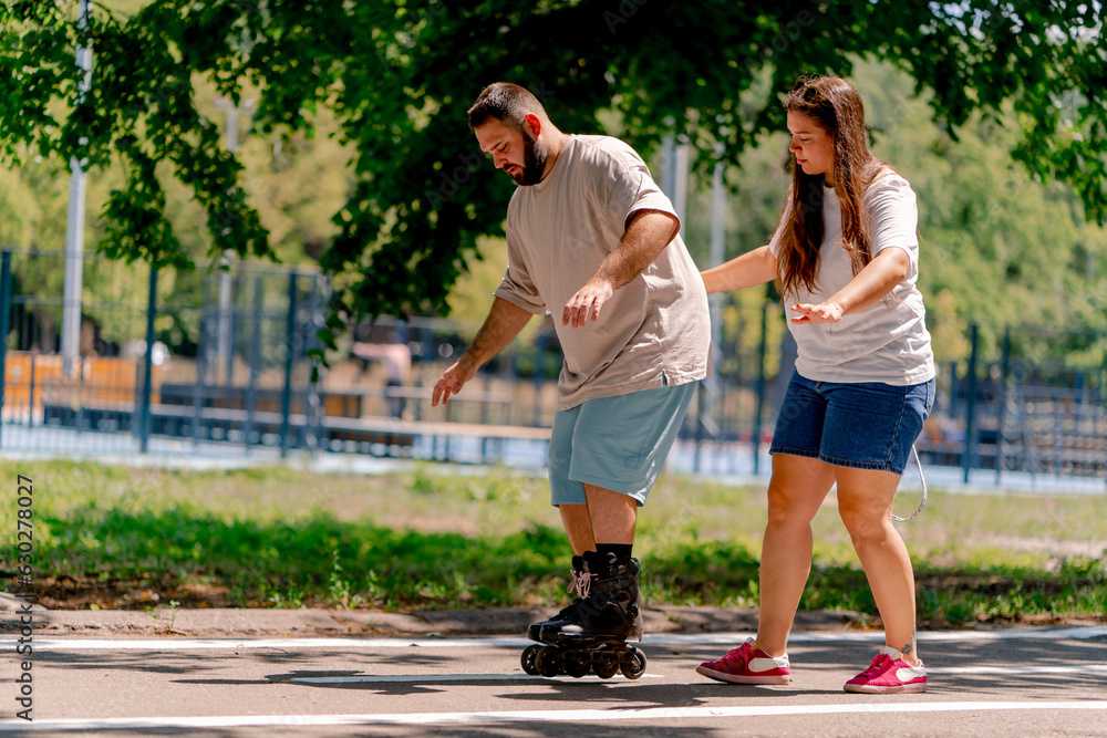 Young couple girl holds hands of the guy who is roller skating and helps him to learn to skate in the park plus size models