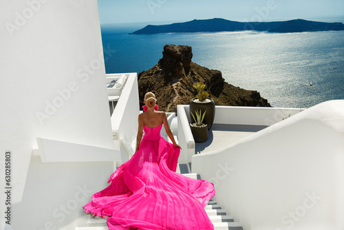 Beautiful elegant girl in long fluttering gown dress is running up the stairs near Skaros Rock in luxury resort in Santorini, Imerovigli. Female model in amazing long dress. photo