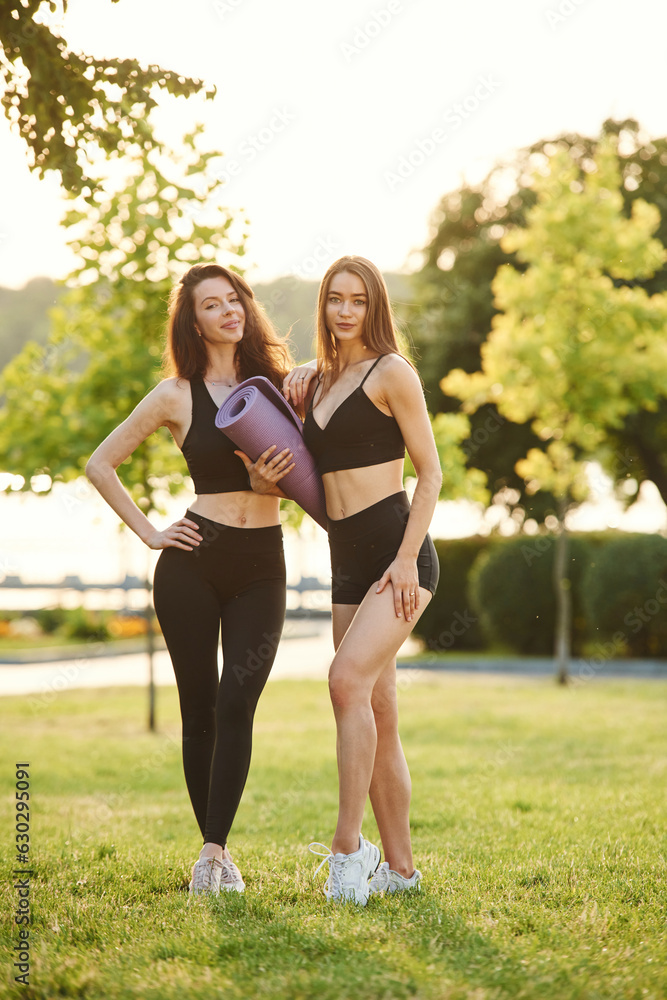 Standing together, with yoga mat. Two women in sport clothes are outdoors on the field Standing together, with yoga mat. Two women in sport clothes are outdoors on the field