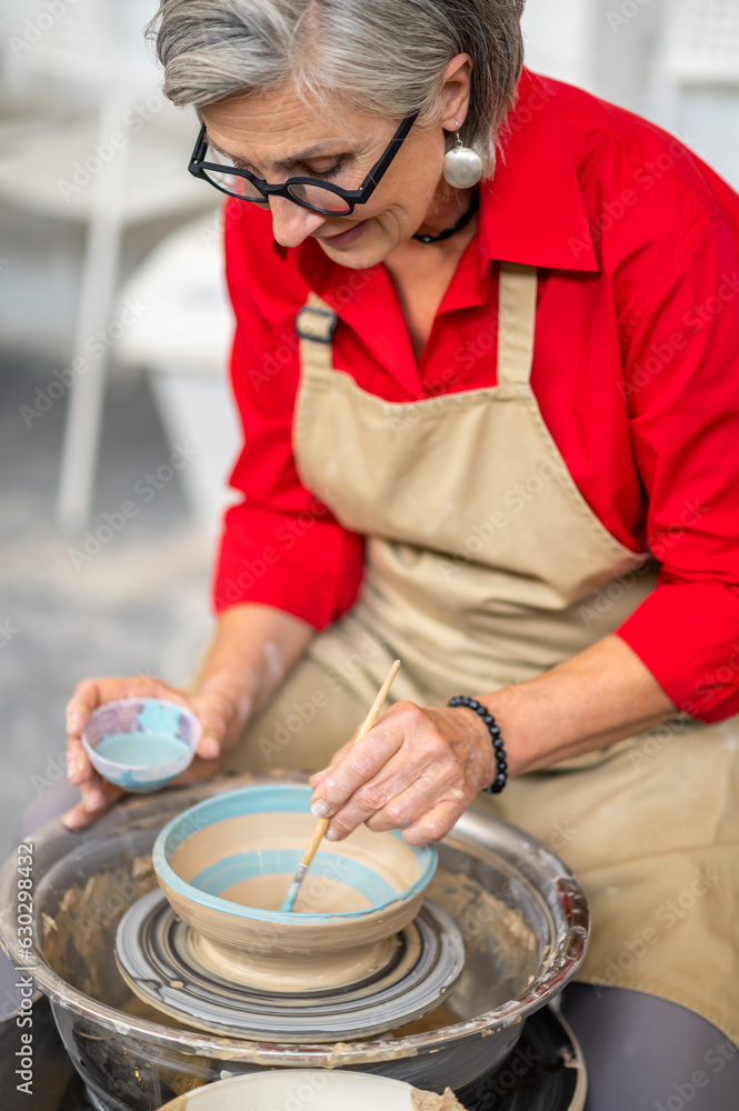 Mature craftswoman painting clay mug blue paint, using pottery wheel and paintbrush.