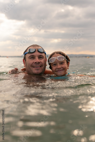 Happy dad swims in the sea with his cute daughter. Girl smiles happily and enjoys a swim in the warm water. The both with water glasses