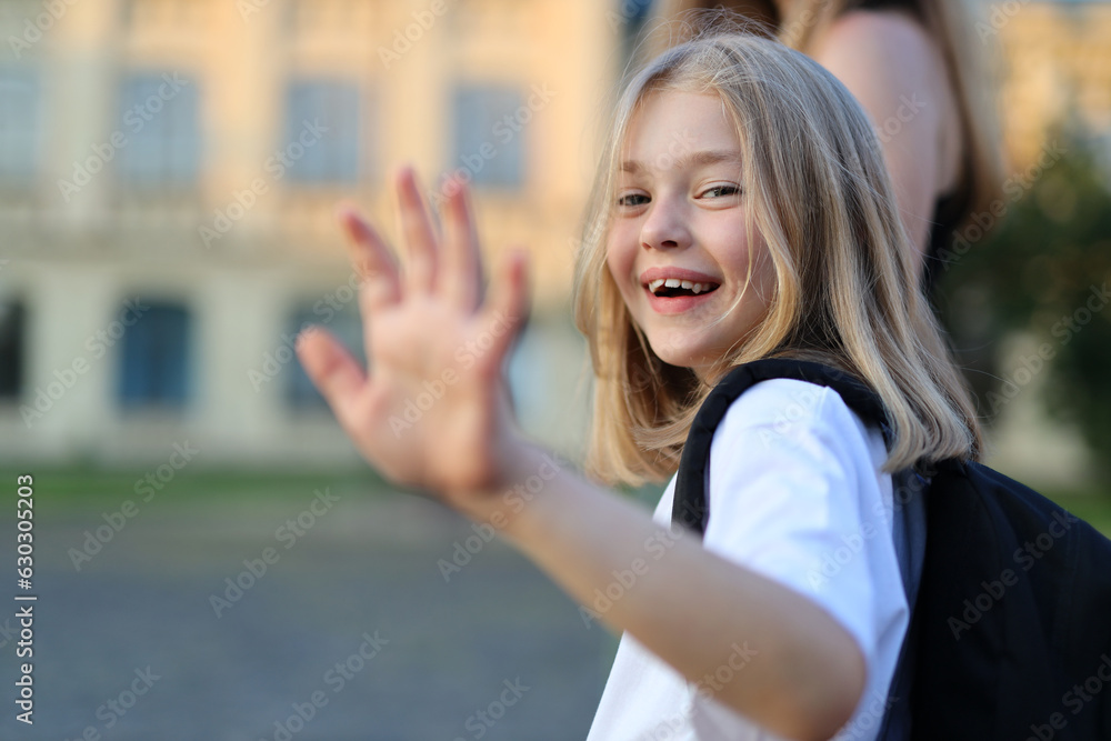 A schoolgirl with a backpack waving in front of a school building. She is a cheerful student walking to school.