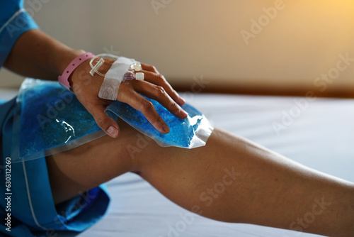 The patient's hand in the hospital with a brine line piercing sitting on the bed holding blue gel ice pack on the knee.
