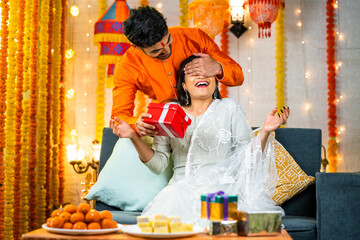 Happy brother by closing eyes giving surprise gift to sister during raksha bandhan festival celebration at home - concept of relationship bonding, togetherness and family ceremony.