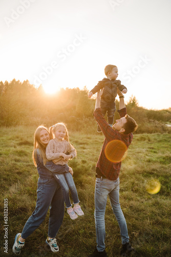 Mother, father, daughter, son walks in grass in spring field at sunset. Childs embrace parents. Family spending time together in nature. Parents hold in hands, throw up happy kids into sky autumn day.