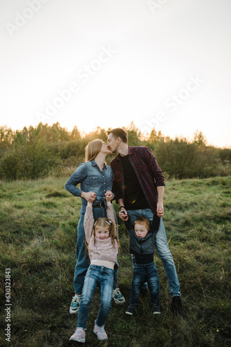 Family spending time together in nature. Parents hold in hands and hugs happy kids in autumn day. Mother, father, daughter, son walks in grass in spring field at sunset. Childs embrace parents.