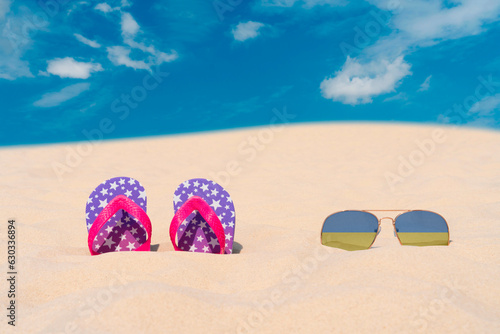 Sunglasses with glasses in the form of the flag of Ukraine and flip-flops lie on the sand against the blue sky. The concept of summer holidays, travel and tourism in Ukraine