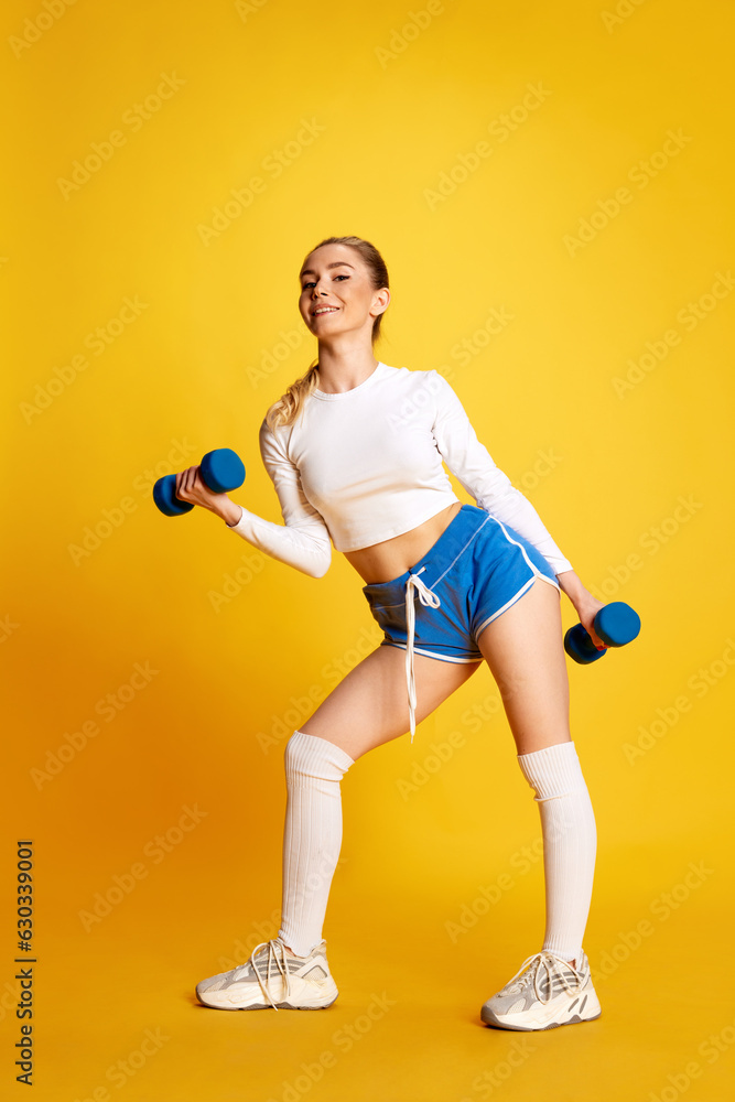 Full lenght image of young woman in training pumping up muscles of hands and legs looking at camera over yellow background. Side view
