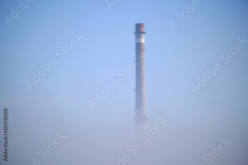 Thick fog from which a factory with a large industrial pipe is visible