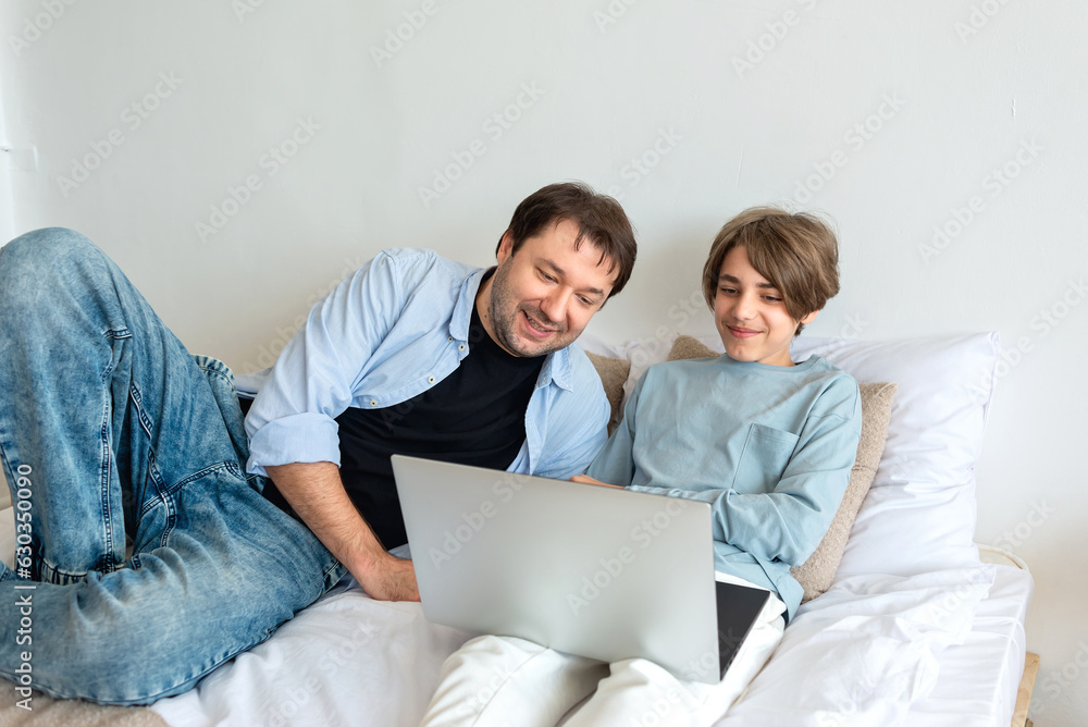 Father and teenager son looking at laptop screen, smile, watching video, communicate online together, in social media, laying on bad at home. Happy family relations, child support, fatherhood concept Father and teenager son looking at laptop screen, smile, watching video, communicate online together, in social media, laying on bad at home. Happy family relations, child support, fatherhood concept