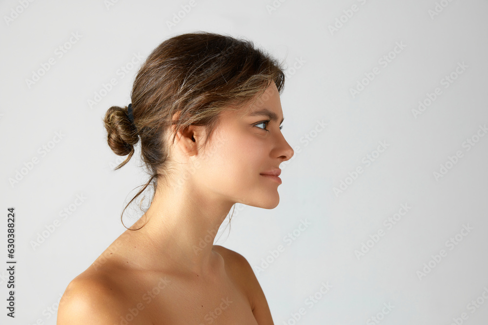 Young woman with hair bun, bare shoulders against grey studio background. Side view portrait. Cosmetological injections. Concept of natural beauty, cosmetology and dermatology, skincare, cosmetics, ad Young woman with hair bun, bare shoulders against grey studio background. Side view portrait. Cosmetological injections. Concept of natural beauty, cosmetology and dermatology, skincare, cosmetics, ad