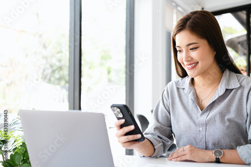 Cheerful woman entrepreneur sitting beside a window and smiling. Businesswoman working in office with a mobile phone in hand. 