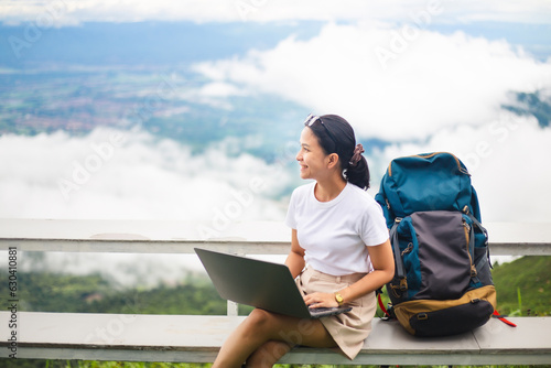 Young smiling woman backpacker using laptop with beautiful sky and mist.  Asian female tourist working at computer while sitting with backpack at resort. Travel and technology concept.