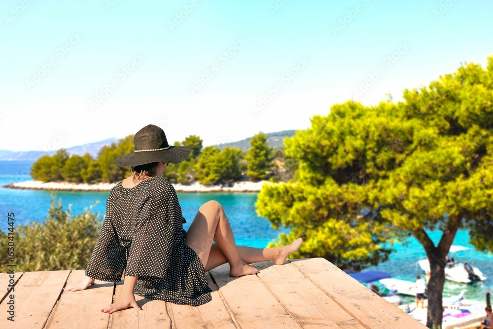 Woman wearing a hat sitting on wooden porch overlooking a calm lake with a few boats in the water